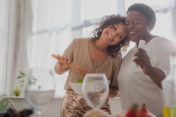 Happy african american woman holding salad near mother with smartphone during thanksgiving celebration — Stock Photo