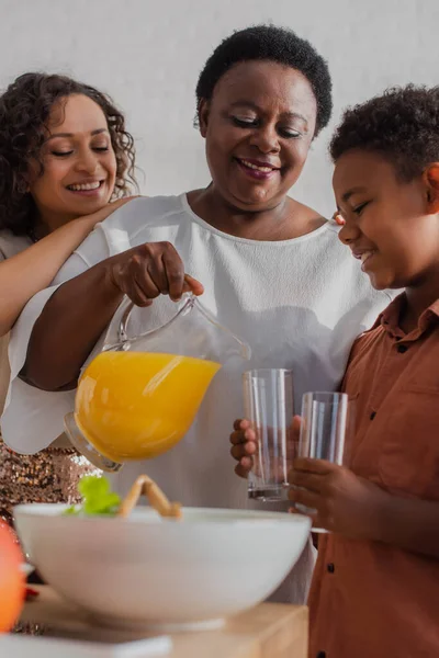 Sorrindo mulher afro-americana derramando suco de laranja perto de neto e filha durante o jantar de ação de graças — Fotografia de Stock