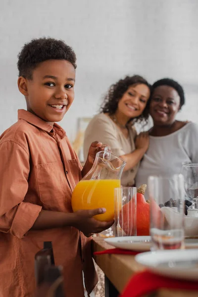 Smiling african american boy holding orange juice near thanksgiving dinner and family at background — Stock Photo