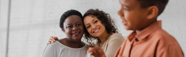 Sorridente donna afro-americana che abbraccia la madre vicino al bambino sfocato a casa, striscione — Foto stock