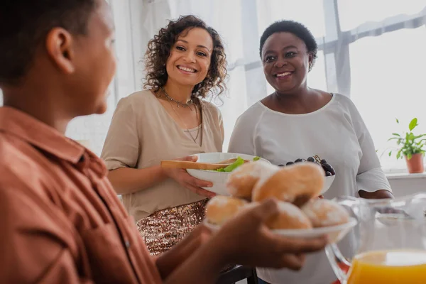 Mulheres africanas americanas sorridentes olhando para menino borrado com pães durante a celebração de ação de graças — Fotografia de Stock