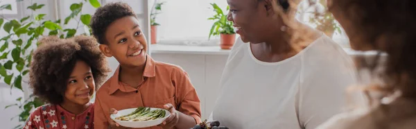 Enfants afro-américains souriants avec des asperges debout près de la mère et la mamie à la maison, bannière — Photo de stock