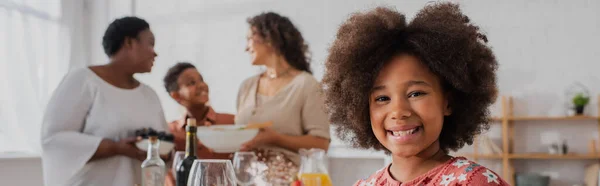 Smiling african american girl looking at camera near blurred family and thanksgiving dinner, banner — Stock Photo