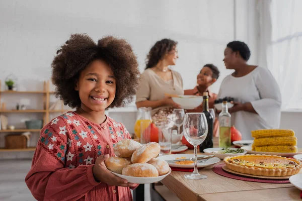 Niño afroamericano sosteniendo bollos cerca de sabrosa cena de acción de gracias y la familia borrosa - foto de stock