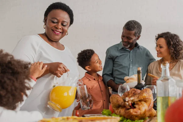 Mulher afro-americana derramando suco de laranja perto da família durante o jantar de ação de graças — Fotografia de Stock