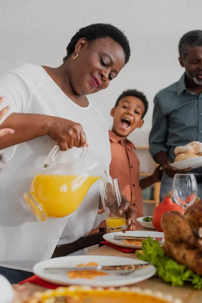 African american granny pouring orange juice near blurred family and thanksgiving dinner — Stock Photo