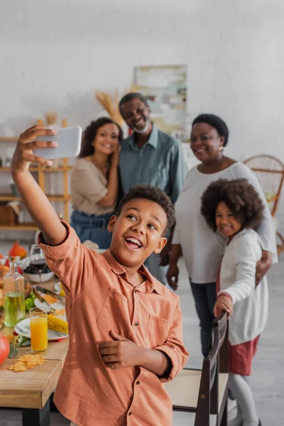 Niño afroamericano tomando selfie con familia borrosa y cena de acción de gracias - foto de stock