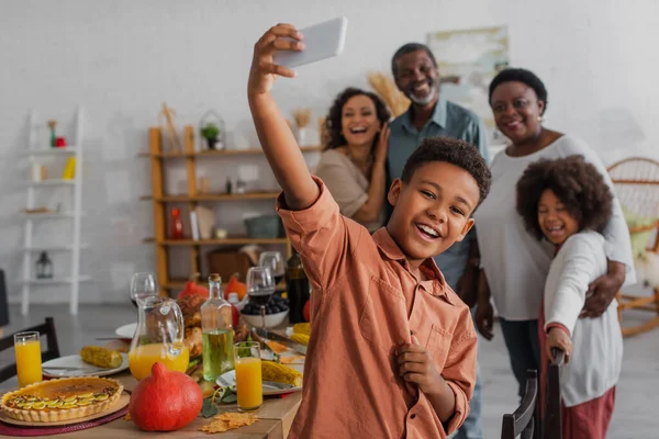 Усміхнена афроамериканська дитина бере селфі на смартфон біля розмитої сім'ї під час вечері подяки — стокове фото
