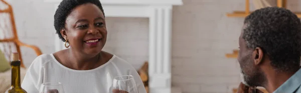 Femme afro-américaine souriante regardant son mari près du vin à la maison, bannière — Photo de stock
