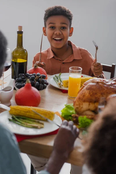 Gai garçon afro-américain tenant des couverts près de délicieux dîner de Thanksgiving et la famille floue — Photo de stock