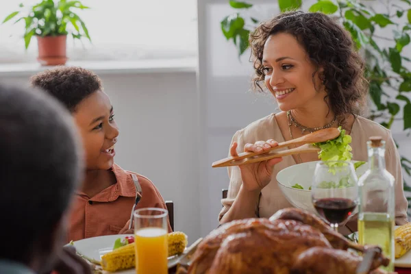 Sonriente mujer afroamericana sosteniendo ensalada cerca de hijo y borrosa cena de acción de gracias - foto de stock