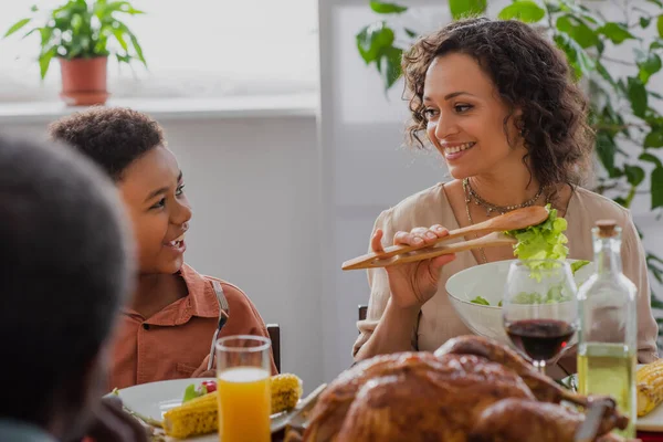 Lächelnde Afroamerikanerin mit Salat in der Nähe ihres Sohnes und verschwommenem Danksagungsdinner — Stockfoto