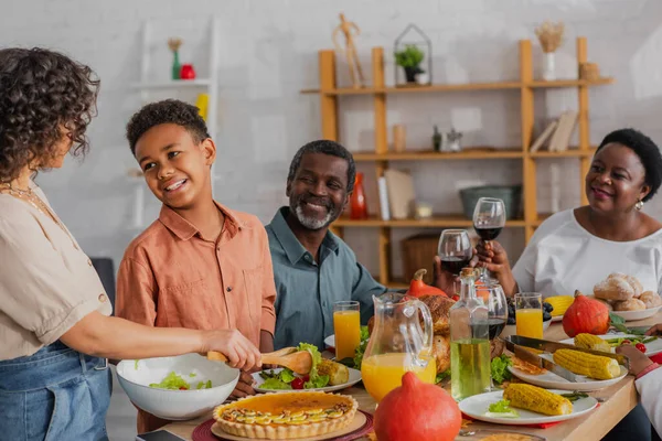 African american boy looking at mother pouring salad during thanksgiving dinner with grandparents — Stock Photo
