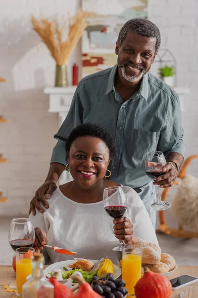 Pareja afroamericana madura con vino mirando la cámara cerca de la cena de Acción de Gracias y el teléfono inteligente - foto de stock