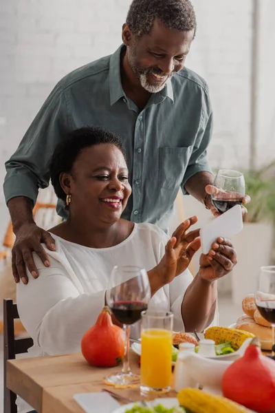 African american woman using smartphone near husband with wine and thanksgiving dinner — Stock Photo
