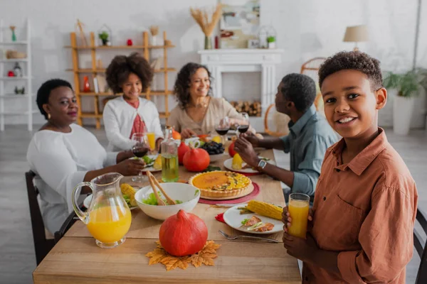Sonriente chico afroamericano sosteniendo jugo de naranja cerca de la familia en la cena de acción de gracias - foto de stock