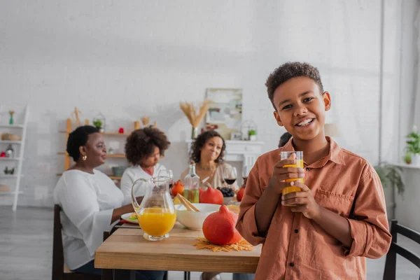 Niño afroamericano sosteniendo jugo de naranja cerca de la familia borrosa durante el día de acción de gracias - foto de stock