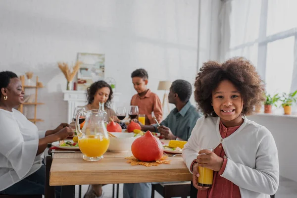 Niño afroamericano con jugo de naranja de pie cerca de la cena decorada de acción de gracias y la familia - foto de stock