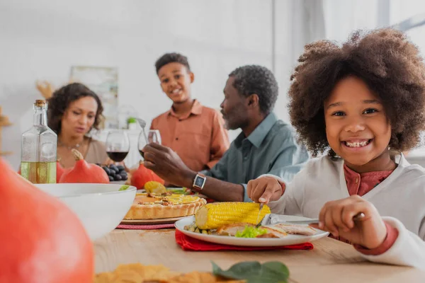 Cheerful african american girl cutting corn near blurred family and thanksgiving dinner — Stock Photo