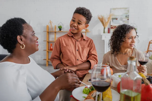 Радостный африканский американец держит за руку бабушку рядом с мамой и столом, подаваемый с праздничным ужином — стоковое фото