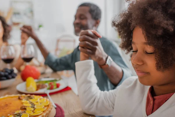 Enfoque selectivo de la chica afroamericana con los ojos cerrados rezando con el abuelo borroso y mamá en la cena de acción de gracias - foto de stock