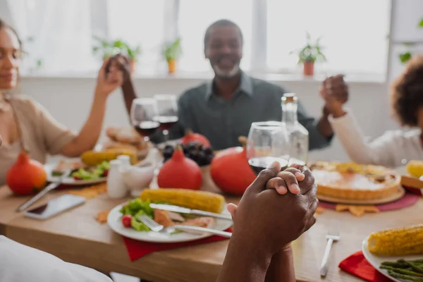 Foco seletivo da mulher afro-americana e menino de mãos dadas enquanto reza com a família turva perto da mesa servida — Fotografia de Stock