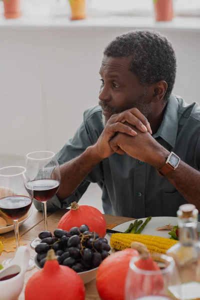 Hombre afroamericano de mediana edad sentado con las manos apretadas en la mesa con cena de acción de gracias - foto de stock