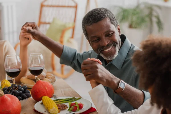 Homme afro-américain joyeux tenant la main avec la famille tout en priant sur le dîner de Thanksgiving — Photo de stock