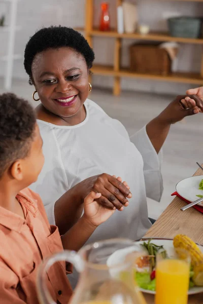 Happy african american woman holding hands with grandson and daughter while praying before thanksgiving dinner — Stock Photo