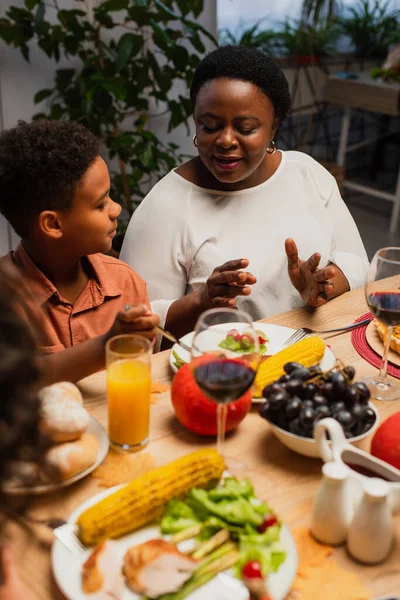 Mature african american woman talking to grandson and blurred daughter near table served with thanksgiving dinner — Stock Photo
