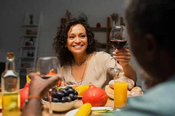 Glückliche afrikanisch-amerikanische Frau prostet sich mit einem Glas Rotwein in der Nähe des verschwommenen Vaters beim Erntedank-Dinner zu — Stockfoto