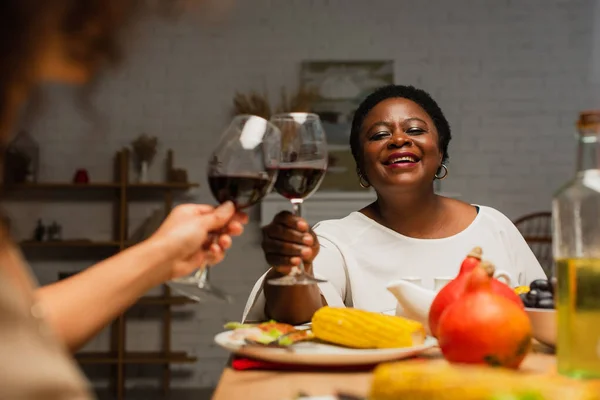 Joyful african american woman clinking glasses of red wine with blurred daughter on thanksgiving dinner — Stock Photo