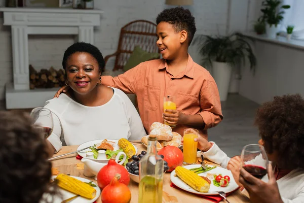 Feliz afroamericano chico con jugo de naranja abrazando abuela durante la cena de acción de gracias con la familia borrosa — Stock Photo