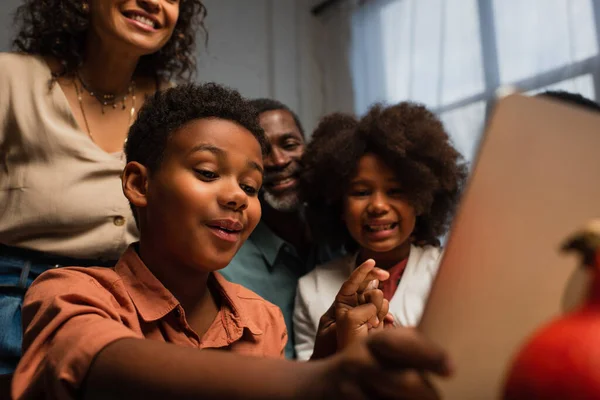 Afroamericano chico gesto durante la videollamada en borrosa portátil cerca de feliz familia - foto de stock