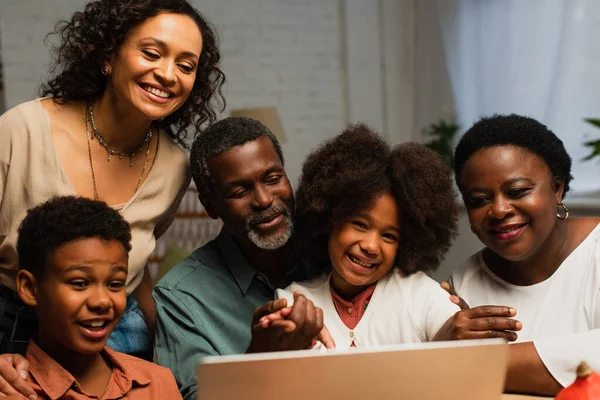 Hombre afroamericano feliz apuntando a la computadora portátil cerca de la familia alegre en el día de acción de gracias - foto de stock