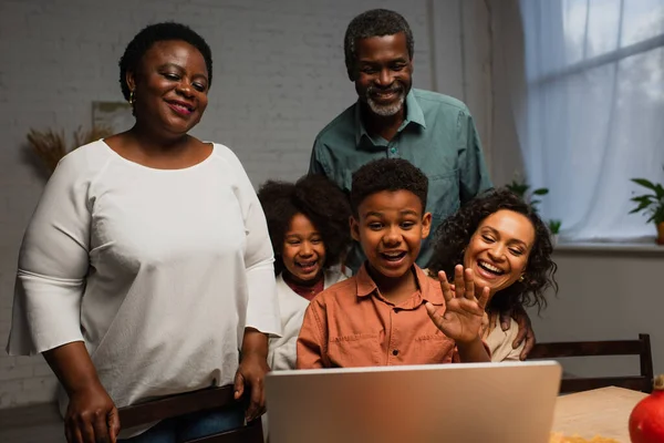 Excited african american boy waving hand during video call near family — Stock Photo