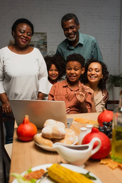 Joyeux garçon afro-américain agitant la main lors d'un appel vidéo près de la famille et servi le dîner — Photo de stock