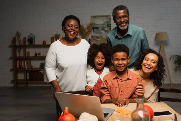Felice famiglia afro-americana sorridente alla fotocamera vicino al computer portatile sul tavolo servito — Foto stock