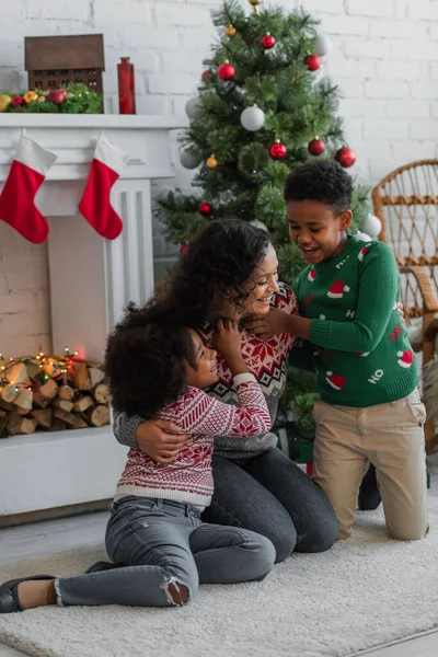 Cheerful african american kids embracing happy mother near christmas tree and decorated fireplace — Stock Photo