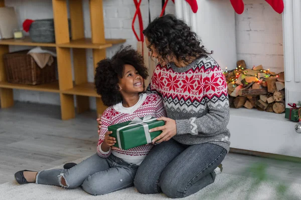 Feliz afroamericano madre e hija sonriendo el uno al otro mientras se sienta cerca de la chimenea decorada con regalo de Navidad - foto de stock