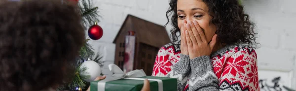 Erstaunt afrikanisch-amerikanische Frau bedeckt Mund mit Händen in der Nähe verschwommene Tochter mit Weihnachtsgeschenk, Banner — Stockfoto