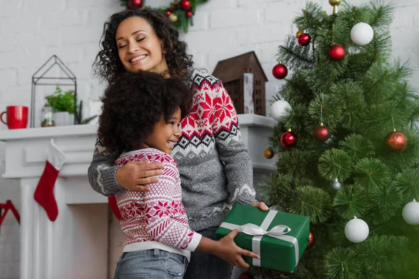 Heureuse femme africaine avec les yeux fermés embrassant fille avec cadeau de Noël — Photo de stock