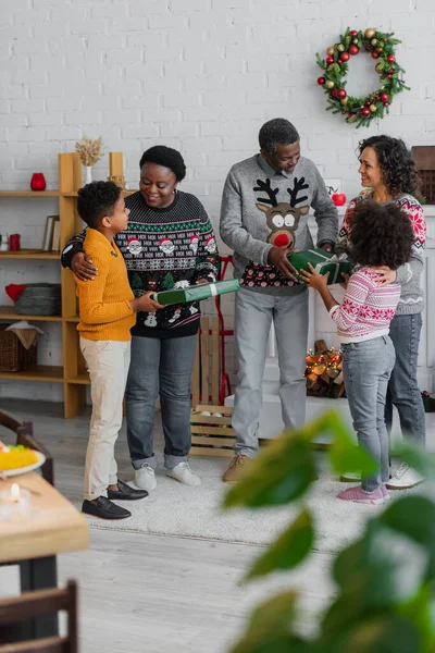 Frère et sœur afro-américain présentant des cadeaux de Noël à des grands-parents heureux près d'une mère souriante — Photo de stock