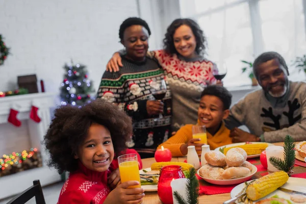 Allegra ragazza afroamericana con bicchiere di succo d'arancia vicino alla cena di Natale e famiglia offuscata — Foto stock