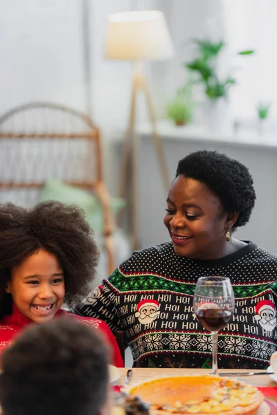 Alegre africana americana chica sonriendo cerca abuelita y borrosa hermano durante la cena de Navidad - foto de stock