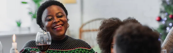 Heureuse femme afro-américaine griller avec du vin rouge tout en célébrant Noël avec des petits-enfants flous, bannière — Photo de stock