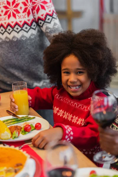 Alegre afroamericana chica con jugo de naranja cerca borrosa abuelos en la cena de Navidad - foto de stock