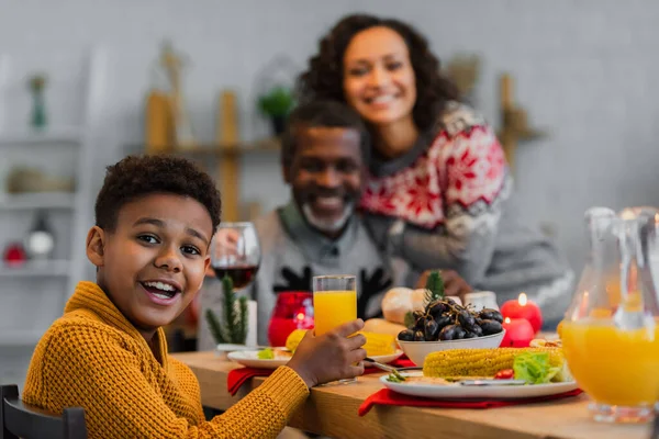 Happy african american boy looking at camera during thanksgiving dinner with blurred family — Stock Photo