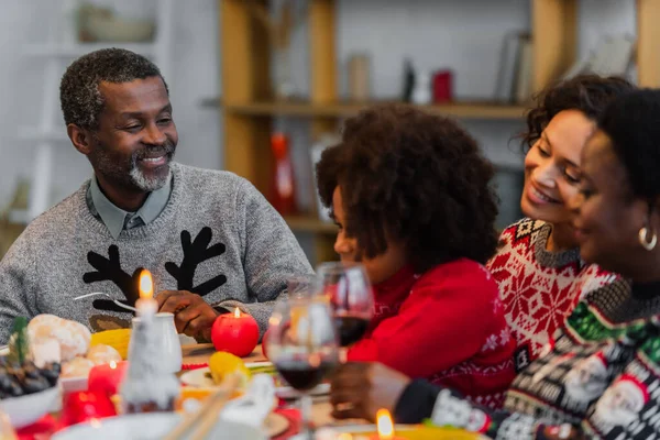 Hombre afroamericano feliz mirando a la nieta cerca de la familia borrosa en la cena de Navidad - foto de stock