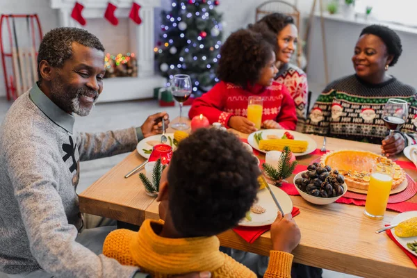 Glücklicher afrikanisch-amerikanischer Mann im Gespräch mit Enkelsohn kurz vor dem Weihnachtsessen mit verschwommener Familie — Stockfoto
